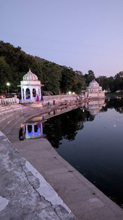 Evening scene of lakeside ghats with small temples lit by colorful lights reflecting on still water while people gather along the steps.
