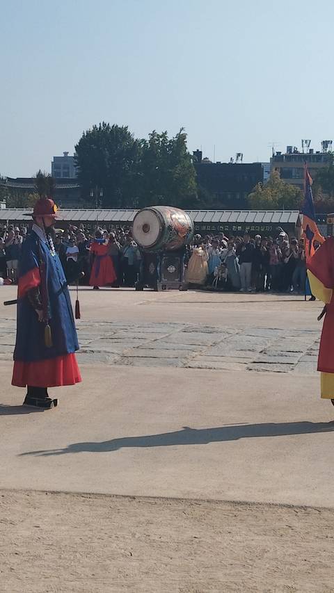 Traditional guard ceremony at a Korean palace courtyard with colorful robes, drum and onlooking crowd.