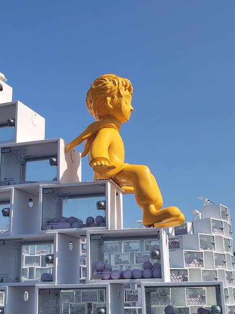 Bright yellow Little Prince statue perched on a modern white structure against a clear blue sky.