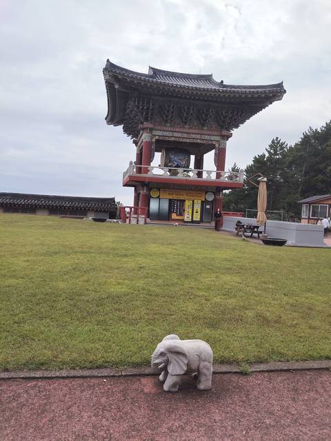 Wide grassy lawn leading to a colorful Buddhist bell tower pavilion on an overcast day.