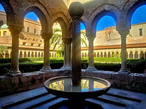 Romanesque cloister with ornate columns, fountain and manicured garden bathed in sunlight.