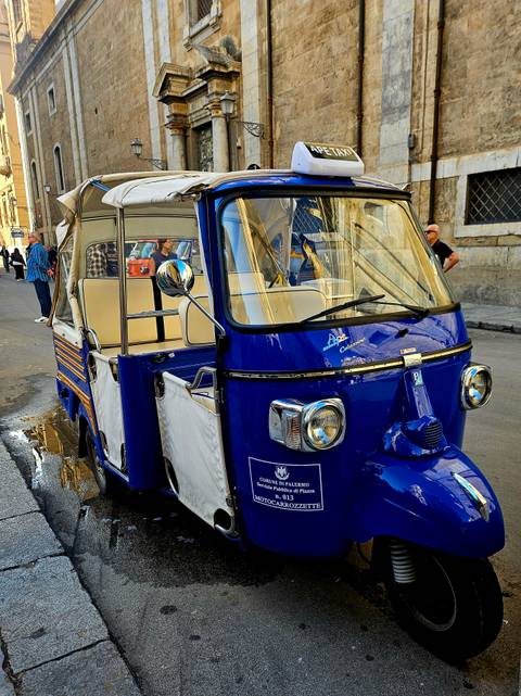 Bright blue three-wheeled tuk-tuk taxi parked on a city street with reflections in a puddle.