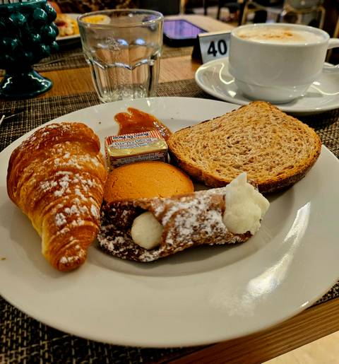 Breakfast plate with croissant, cannoli, biscuit, jam and slice of bread on white dish.