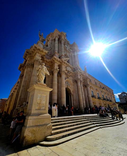 Grand baroque cathedral in Syracuse’s Piazza Duomo lit by intense sun with marble statue in foreground and tourists below.