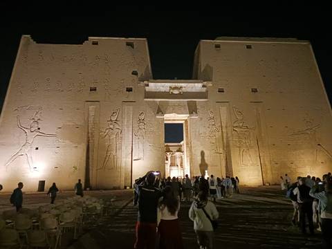 Night tourists entering Edfu Temple façade lit with warm lights revealing carved hieroglyphs.