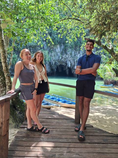Three travellers posing in front of the entrance to an underground river cave with emerald water.