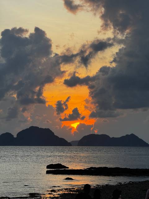 Dramatic orange and yellow sunset breaking through dark clouds above jagged island peaks.