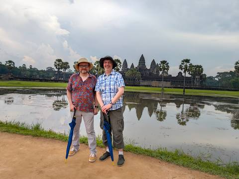 Two tourists stand with umbrellas by the reflective pond opposite Angkor Wat’s iconic towers