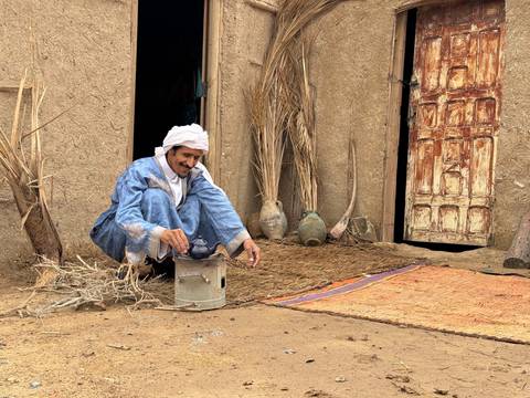 Berber man squats outside an adobe home brewing tea on a small metal stove.