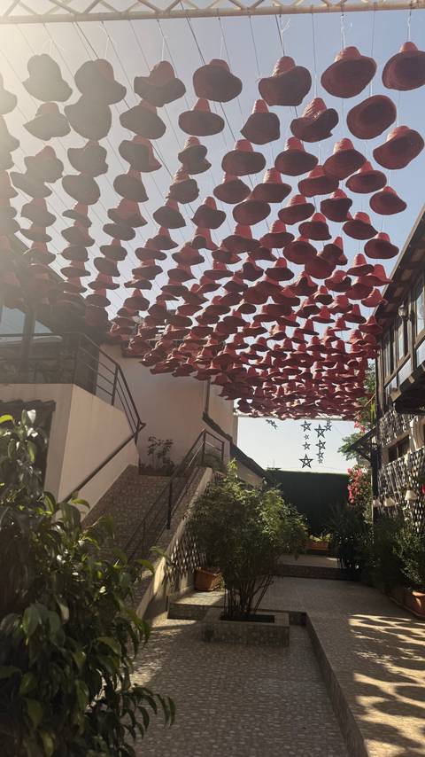 A narrow white-walled alley with a staircase and dozens of red straw hats suspended overhead, lit by bright midday sun.