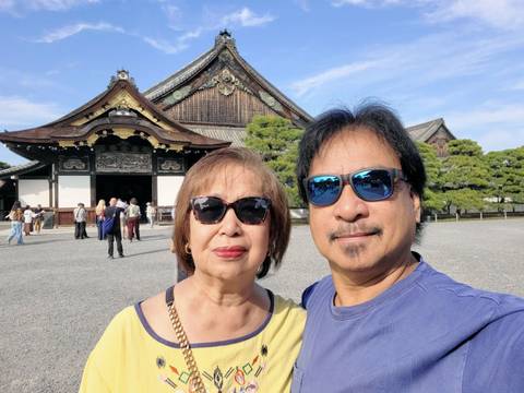 Couple selfie with the ornately roofed Nijo Castle gate and courtyard behind them in bright sunlight.