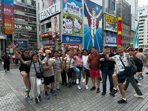 Cheerful tour group poses in Osaka’s neon-lit Dotonbori street with famous billboards and signs.