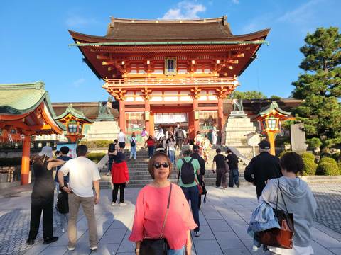 Visitor stands before the vivid orange entrance gate of Fushimi Inari Shrine while crowds ascend the steps.