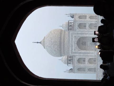 Crowds photograph the Taj Mahal framed by a dark archway in early morning haze.