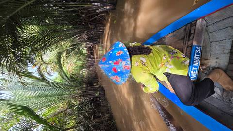 Woman in conical hat paddles a wooden boat through narrow, palm-shaded canal in the Mekong Delta