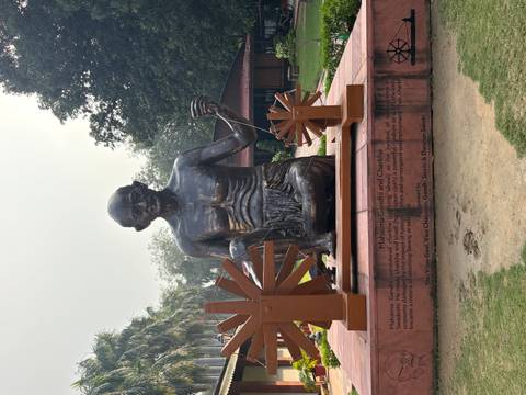 Large bronze statue of Mahatma Gandhi spinning a charkha in a serene garden setting.