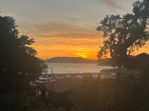 Fiery sunset over a tranquil bay framed by silhouetted trees and boats.