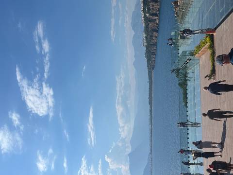 Visitors walk across a glass viewpoint platform overlooking the Mediterranean and cliffs.