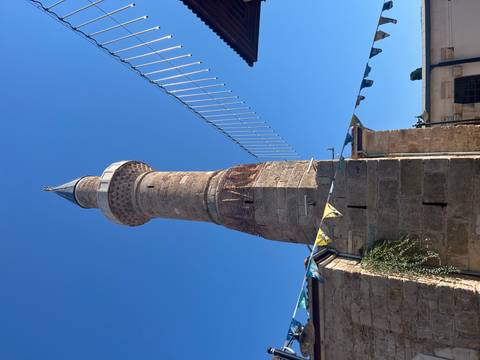 Stone minaret of a historic mosque rising against a clear blue sky with festive bunting below.