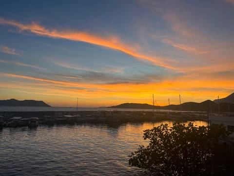 Colorful sunset over a marina with yachts and distant hills reflecting on calm water.