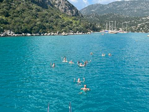 Swimmers enjoying bright turquoise waters in a sheltered bay with sailboats in the distance.