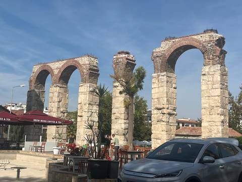 Three surviving arches of an ancient aqueduct beside a modern café terrace and parked car.