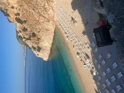 Idyllic sandy cove with turquoise water, lined with neat rows of white sun loungers beneath steep cliffs.