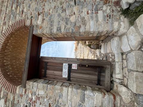 Wooden gate within a stone wall opening to ancient ruins, with a small notice sign attached.