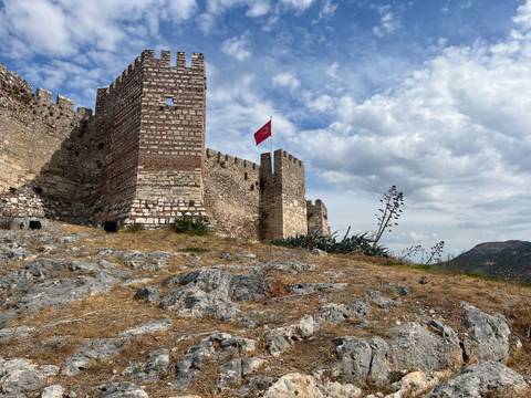 Hilltop stone castle with Turkish flag flying, set against a partly cloudy sky.