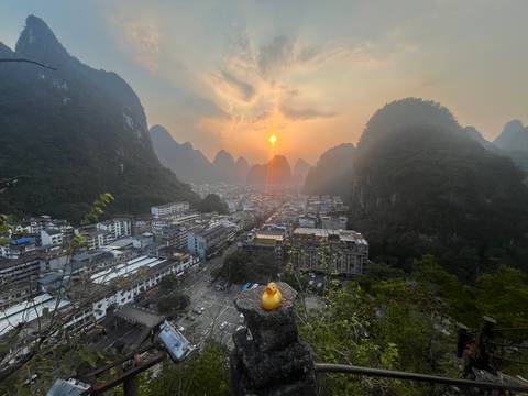 Sunset over a Chinese town nestled among dramatic karst peaks, with a rubber duck ornament in the foreground.