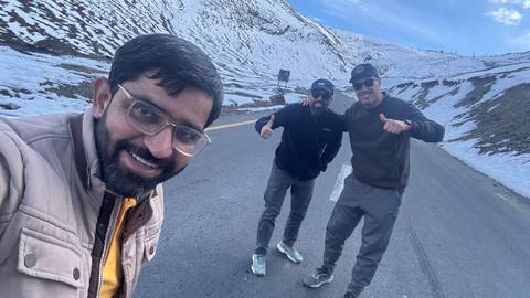 Selfie of three men on a snow-covered mountain road giving thumbs-up under blue sky.