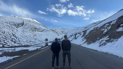 Two men stand on a deserted mountain road flanked by snowy slopes and a blue sky.