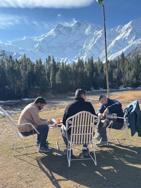 Three men sit outdoors enjoying tea with snow-capped peaks and pine trees in the background.