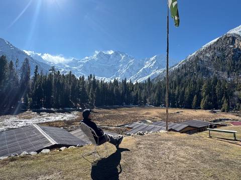 A lone traveler relaxes on a chair facing snow-covered peaks and dense pine forest at Fairy Meadows.