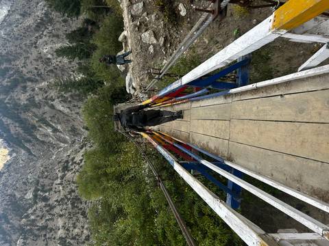 A man crosses a narrow wooden suspension bridge over a lush gorge while another person watches.