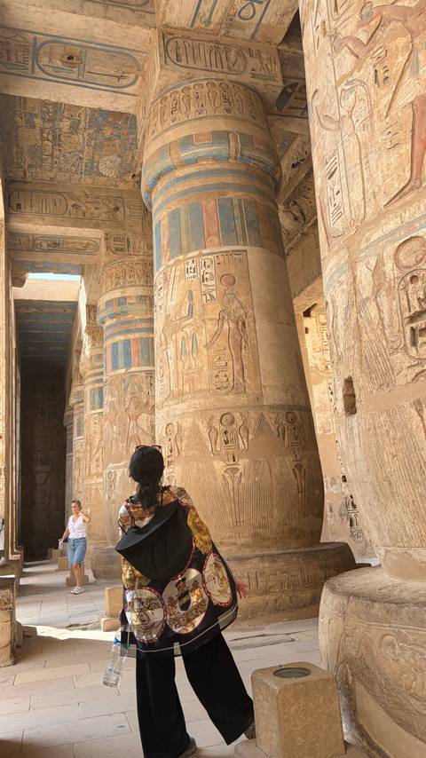 Visitor gazes at hieroglyphic reliefs on massive sandstone columns inside an ancient Egyptian temple.