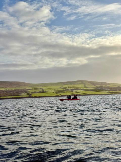 Two kayakers paddle across choppy water with patchwork green fields rolling behind them.