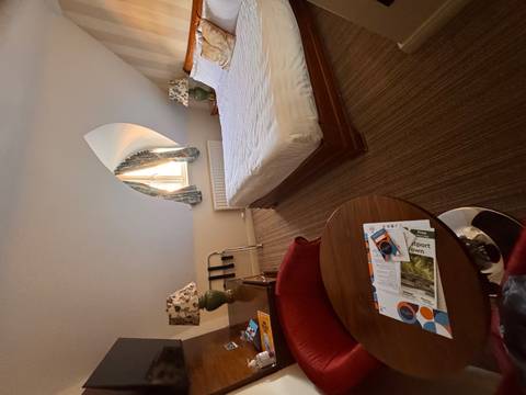 Bright hotel room with sloped ceiling, desk and neatly made bed beneath an arched window.