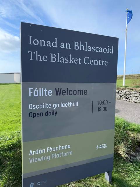 Close-up of bilingual sign for The Blasket Centre on a grassy coastal landscape.