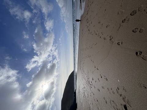 Wide sandy beach with scattered footprints leading to waves under a bright blue sky and scattered clouds.