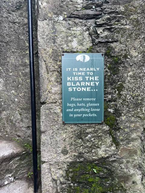 Green sign on stone wall advising visitors about kissing the Blarney Stone.