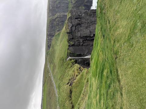Green waterfall plunges over dramatic dark cliffs topped with windswept grass under gray sky.