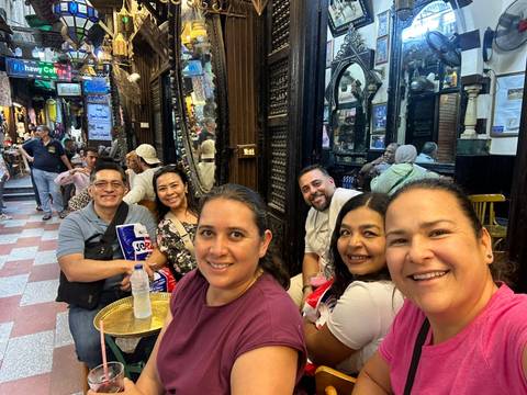 Friends relax with drinks inside a bustling traditional bazaar café lined with ornate woodwork.