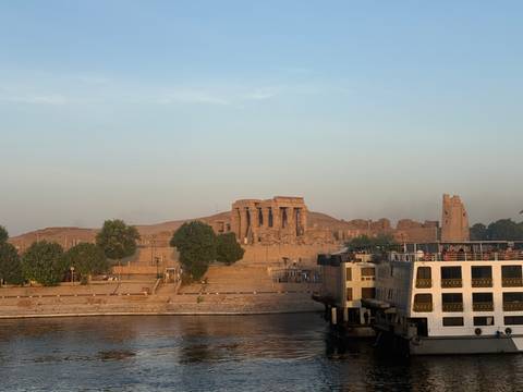 Ancient Kom Ombo Temple rises along the Nile riverbank at dawn with a cruise boat nearby.