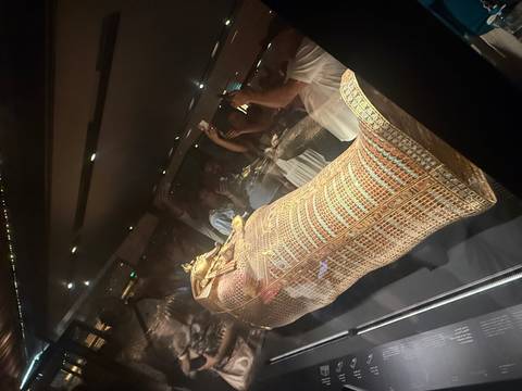 Golden mummy sarcophagus on display inside a dimly lit museum draws photographing visitors.
