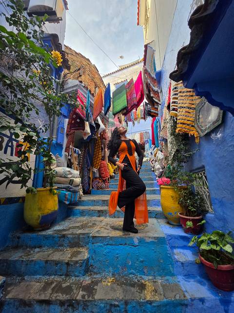 Woman posing confidently on bright blue stairs draped with colorful textiles in a Moroccan alley.