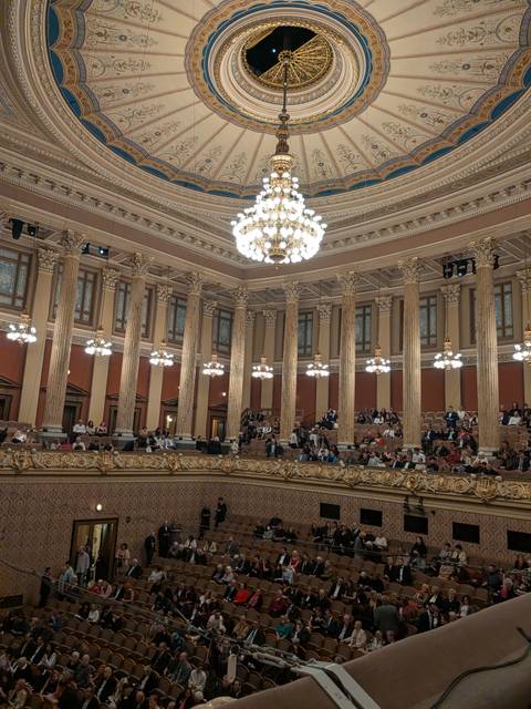 Elegant concert hall interior with ornate columns, chandeliers and seated audience.
