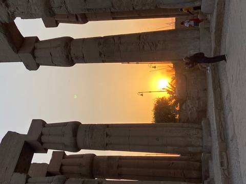 Silhouetted visitor photographing sunset between towering columns of Luxor Temple.