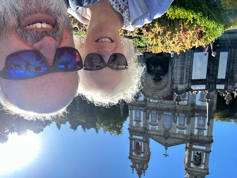 Older couple taking a sunny selfie in front of the ornate twin-towered sanctuary building and formal gardens