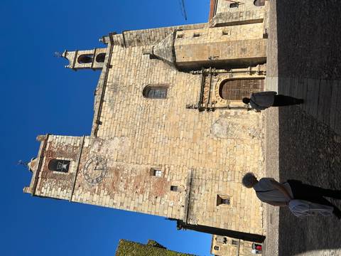 Stone church façade with clock tower against a blue sky while travellers walk across the courtyard